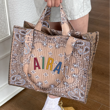 Görseli Galeri görüntüleyiciye yükleyin, A person holds a large tan quilted tote bag with a paisley print and colorful embroidered letters spelling 'AIRA' on the front, shown in an indoor setting with wood flooring.
