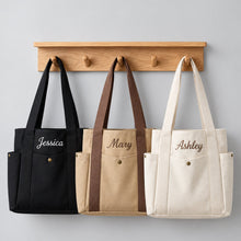 Carica l'immagine nel visualizzatore di Gallery, Three personalized canvas tote bags in black, beige, and cream hang from a wooden coat rack against a light gray wall. The bags feature embroidered names: Jessica, Mary, and Ashley.
