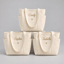 Carica l'immagine nel visualizzatore di Gallery, Three cream canvas tote bags with personalized names (Emily, Sophia, Isabella) in gold script on the front pockets, styled on wooden wedding chairs with white flowers and lace tablecloth.
