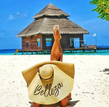 Load image into Gallery viewer, Person relaxing on beach wearing oversized straw sun hat with Belleza text near tropical water bungalow
