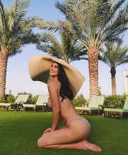 Load image into Gallery viewer, Woman wearing oversized straw sun hat kneeling on grass with palm trees and lounge chairs in background
