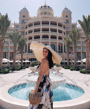 Load image into Gallery viewer, Woman wearing oversized straw sun hat and floral dress standing by fountain in front of elegant building
