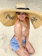 Load image into Gallery viewer, Woman wearing oversized straw sun hat with wide brim and black ribbon sitting on sandy beach
