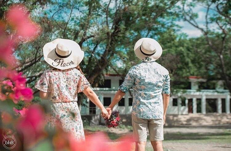 Couple wearing custom prenuptial hat set holding hands outdoors near green trees