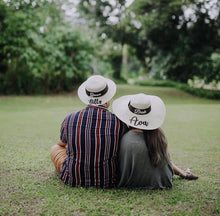Cargar imagen en el visor de la galería, Couple sitting on grass wearing custom prenuptial hat set with personalized names from Travellers Hat Boutique
